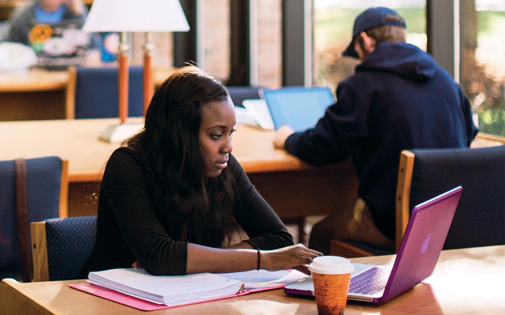 Female student looking a laptop screen while sitting at a wooden table in Miller Library at Cornerstone University