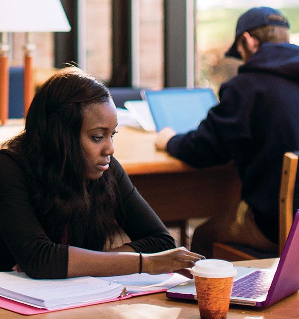 Female student looking a laptop screen while sitting at a wooden table in Miller Library at Cornerstone University