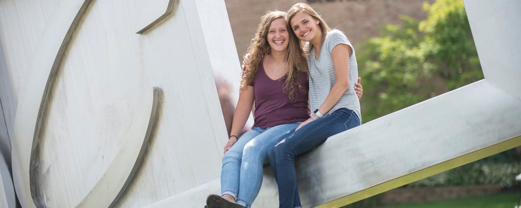 Two female students sitting in the opening of the logo sculpture located outside at Cornerstone University