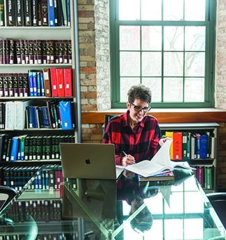 Female adult business student working on her laptop and with a book at a long table with bookshelves in the background