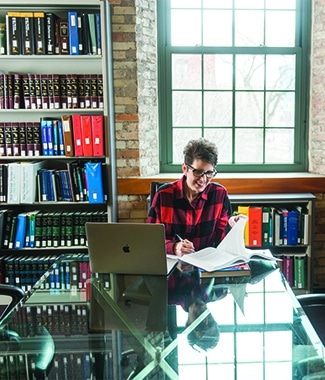 Female adult business student working on her laptop and with a book at a long table with bookshelves in the background