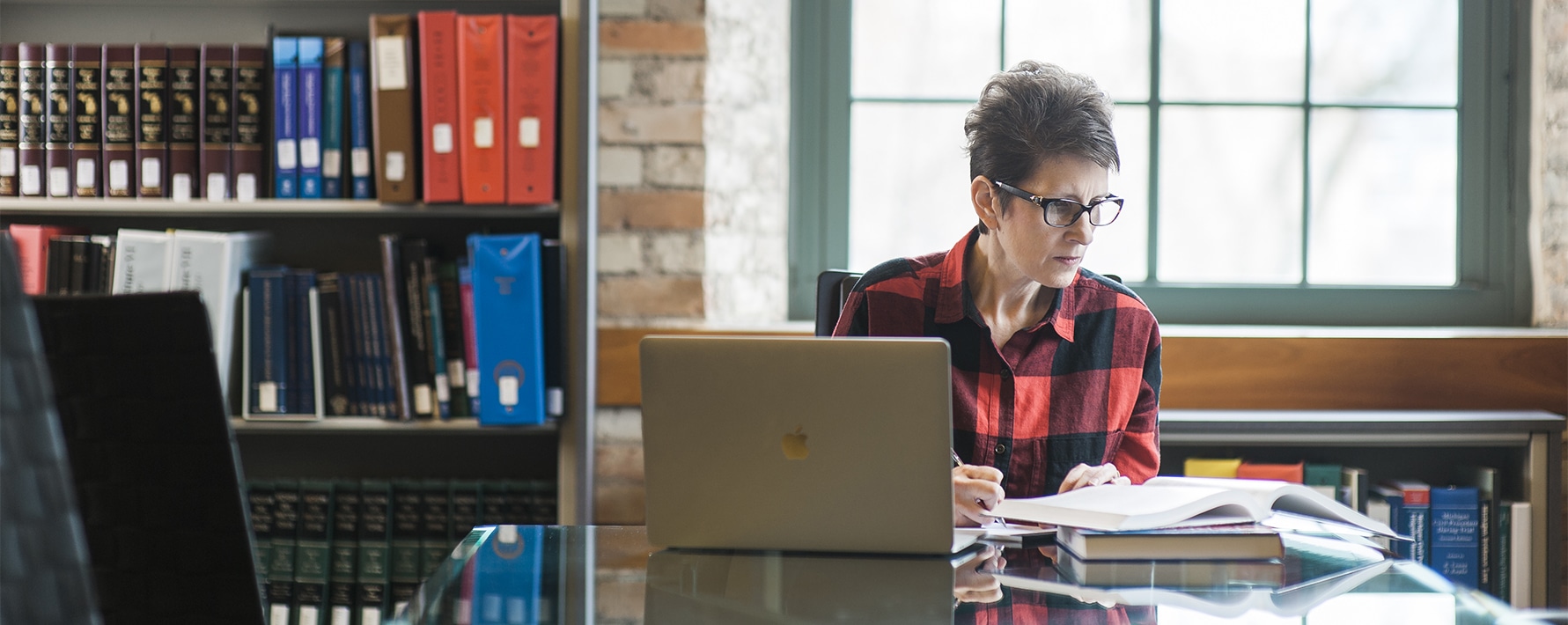 Female adult student working on a laptop and open book at a long table with bookshelves and a window in the background