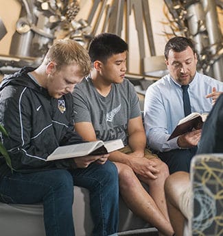 Male professor leading a bible study with male college students in lobby with metal sculpture