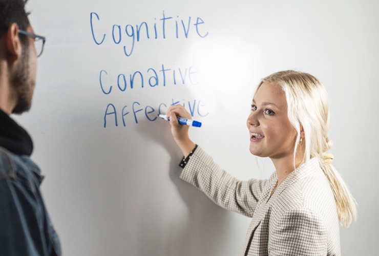 Female in business blazer writing on whiteboard while a male student stands nearby