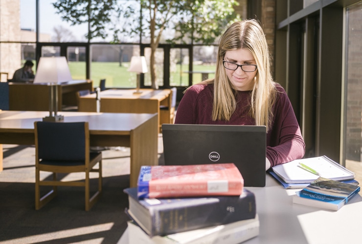 Female college student typing on a laptop at a table by a window in Miller Library at Cornerstone University