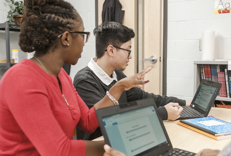 Female graduate student from Cornerstone University teaching at a table with one student with an open laptop