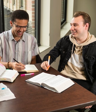 Male students sitting in front of a table with open Bibles while holding pens