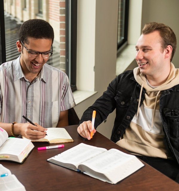 Male students sitting in front of a table with open Bibles while holding pens