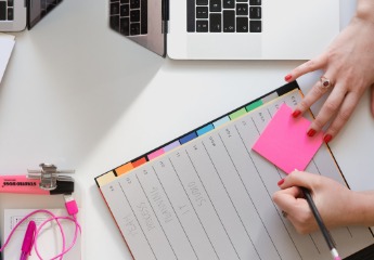 Person writing on a sticky note with an organizational planner and laptop in the background