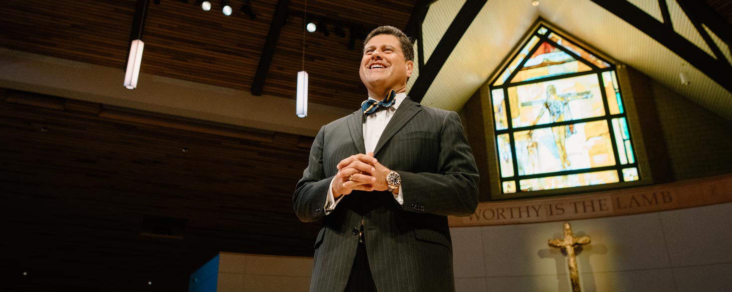 Gerson Moreno-Riaño gazing into the distance while standing on the stage of Christ Chapel at Cornerstone University with a stained glass window and cross in the background
