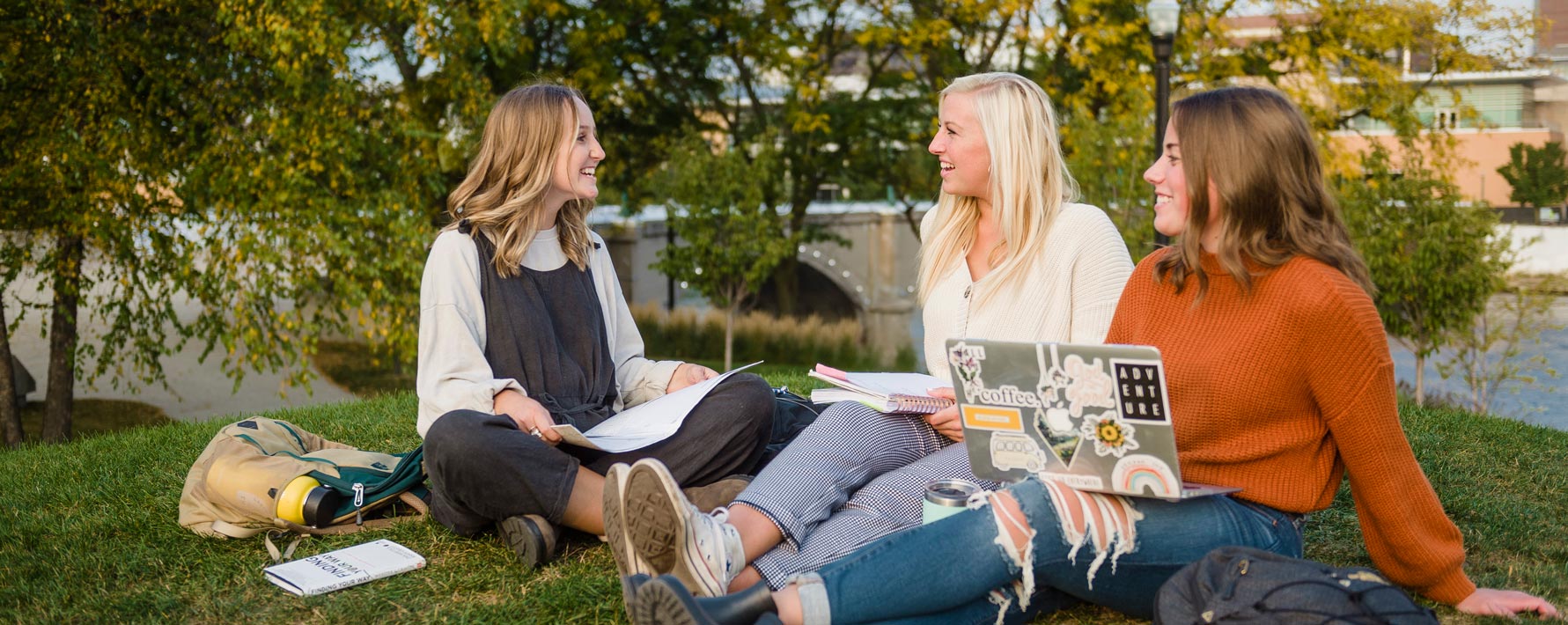 Three female college students studying on grassy knoll in Grand Rapids with a bridge in the background