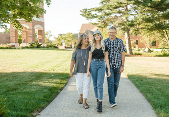 Female college student walking with mother and father on college campus sidewalk on a sunny day