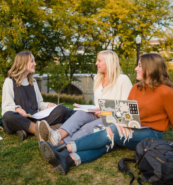 Three female college students studying on grassy knoll in Grand Rapids with a bridge in the background