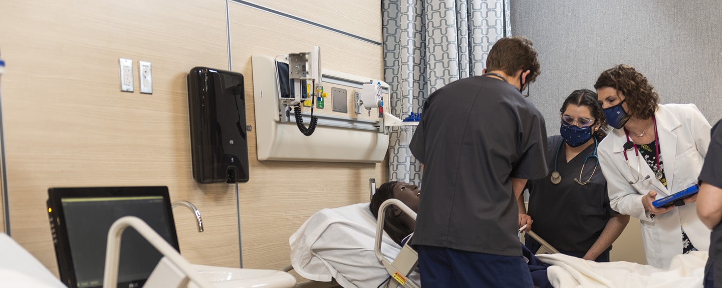 Female nursing instructor oversees two students training on a mannequin in simulated hospital setting