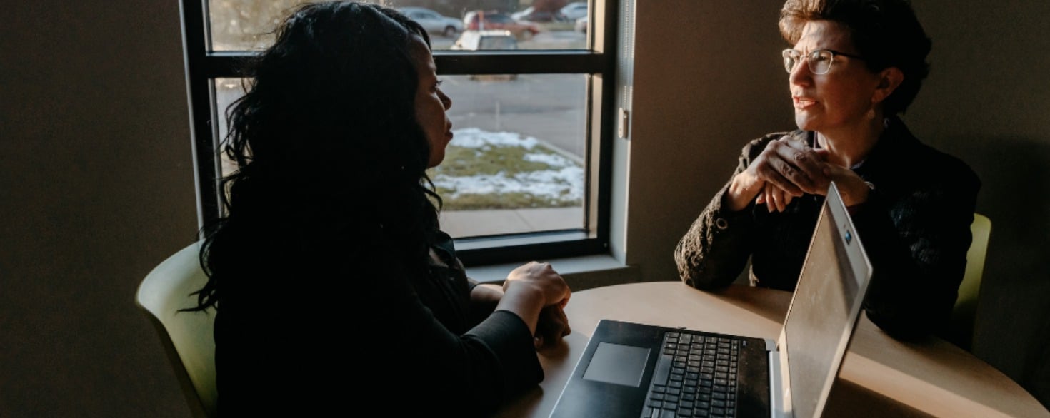 Two women talking with each other while sitting at a table with an open laptop