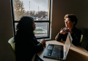 Two women talking with each other while sitting at a table with an open laptop