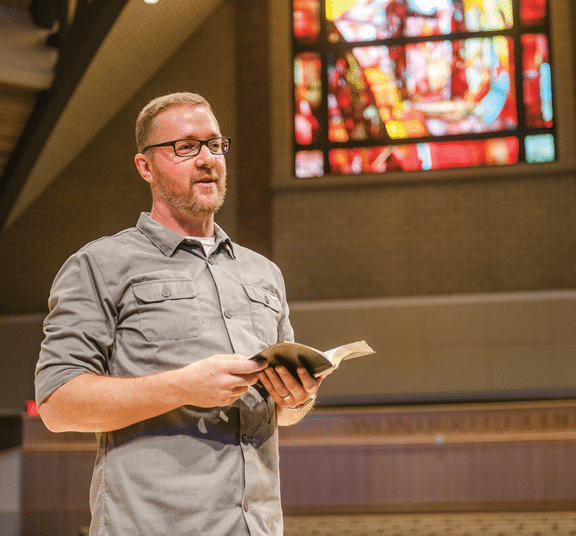 Male speaking in chapel sanctuary with stained glass window while holding an open Bible