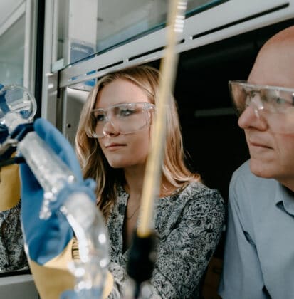 Female student pouring chemicals from a glass beaker in a lab while wearing gloves and safety glasses as Dr. Ned Keller watches