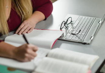 female student at Cornerstone University writing in a notebook with a textbook and laptop nearby