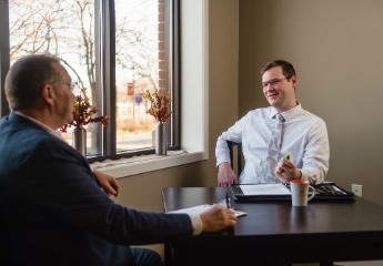 Male employee sitting at a table talking with another male employee.