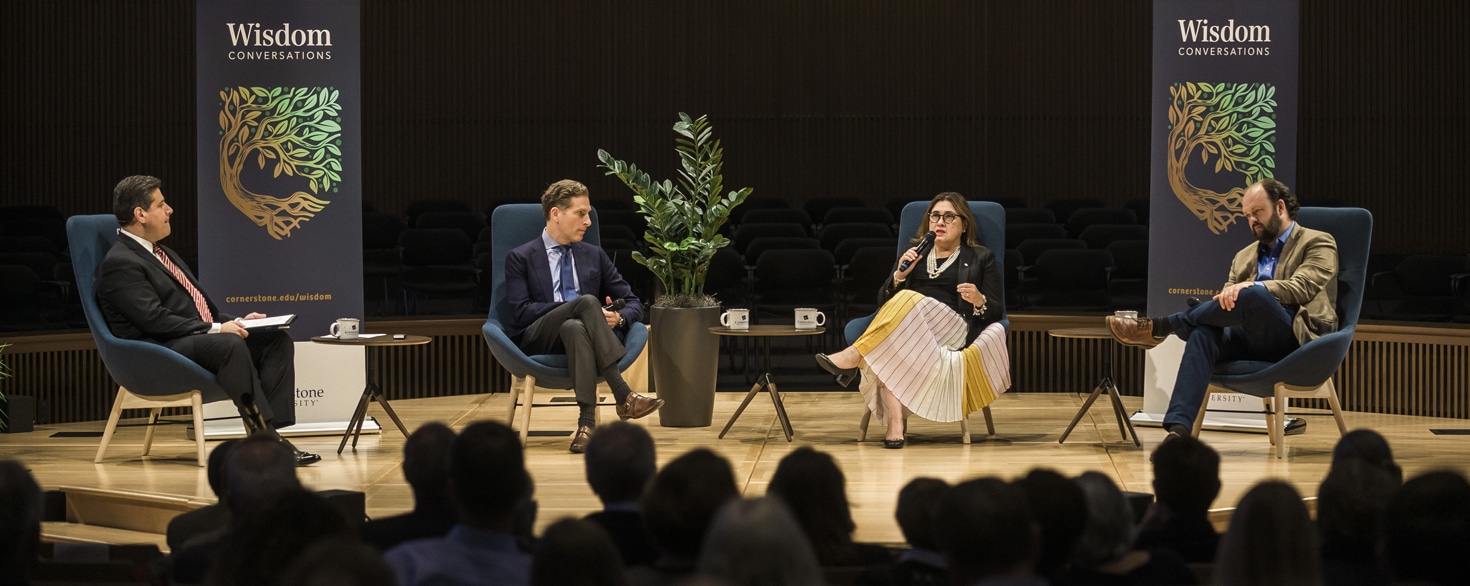 Noah Feldman, Mary Ann Gomez Orta, Gerson Moreno-Riaño and Ross Douthat sitting in chairs on the stage in Christ Chapel at Cornerstone University in front of banners that say Wisdom Conversations