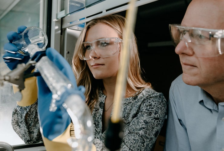 Female college student pouring a beaker while a male professor looks on