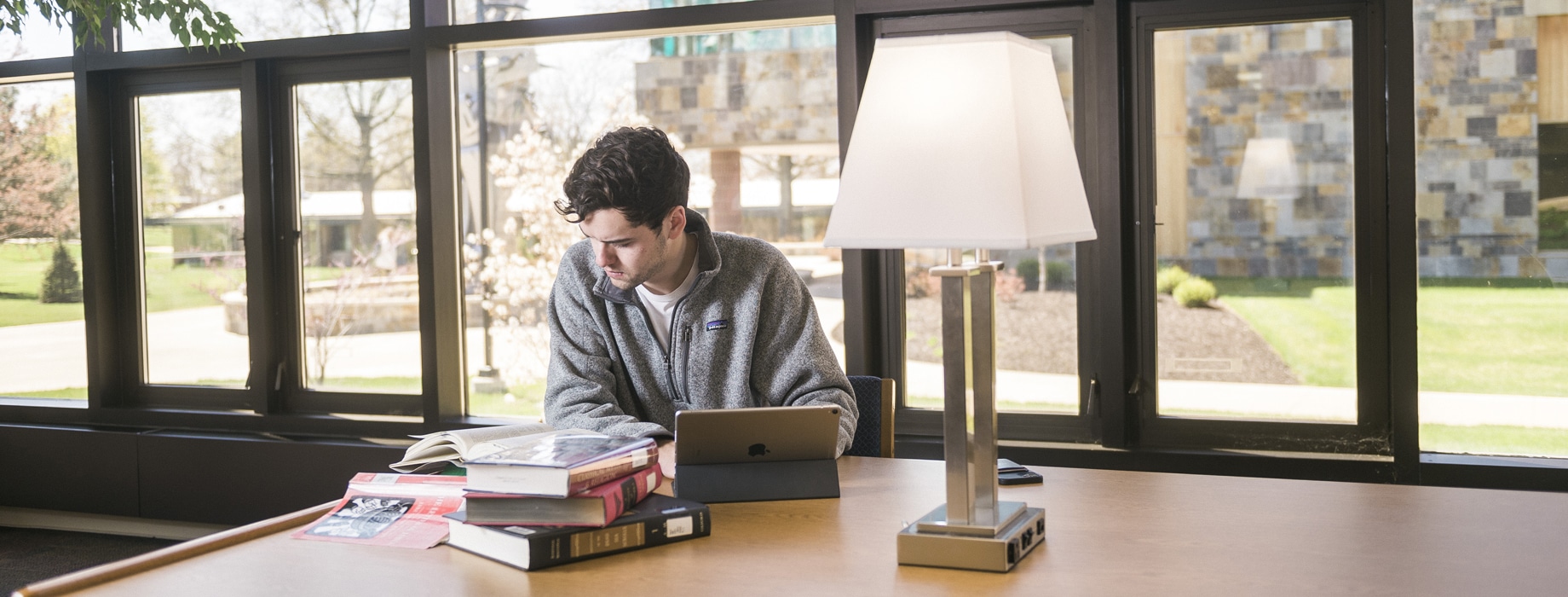 Male student studying at a wooden table with a lamp and stack of hardcover books in Miller Library at Cornerstone University