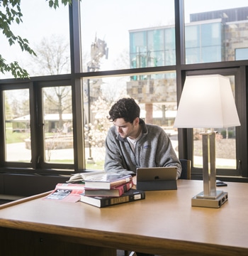 Male student studying at a wooden table with a lamp and stack of hardcover books in Miller Library at Cornerstone University