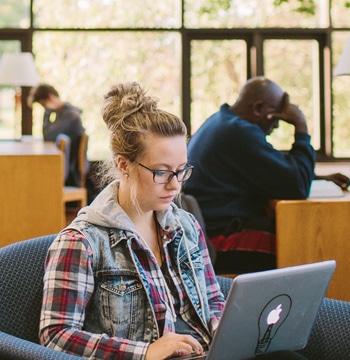 Female student sitting in a chair while typing on a laptop in Miller Library at Cornerstone University