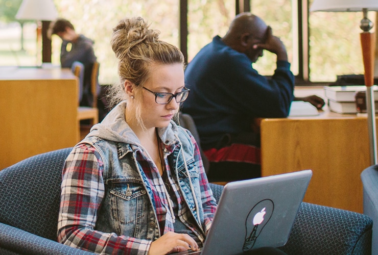 Female student sitting in a chair while typing on a laptop in Miller Library at Cornerstone University