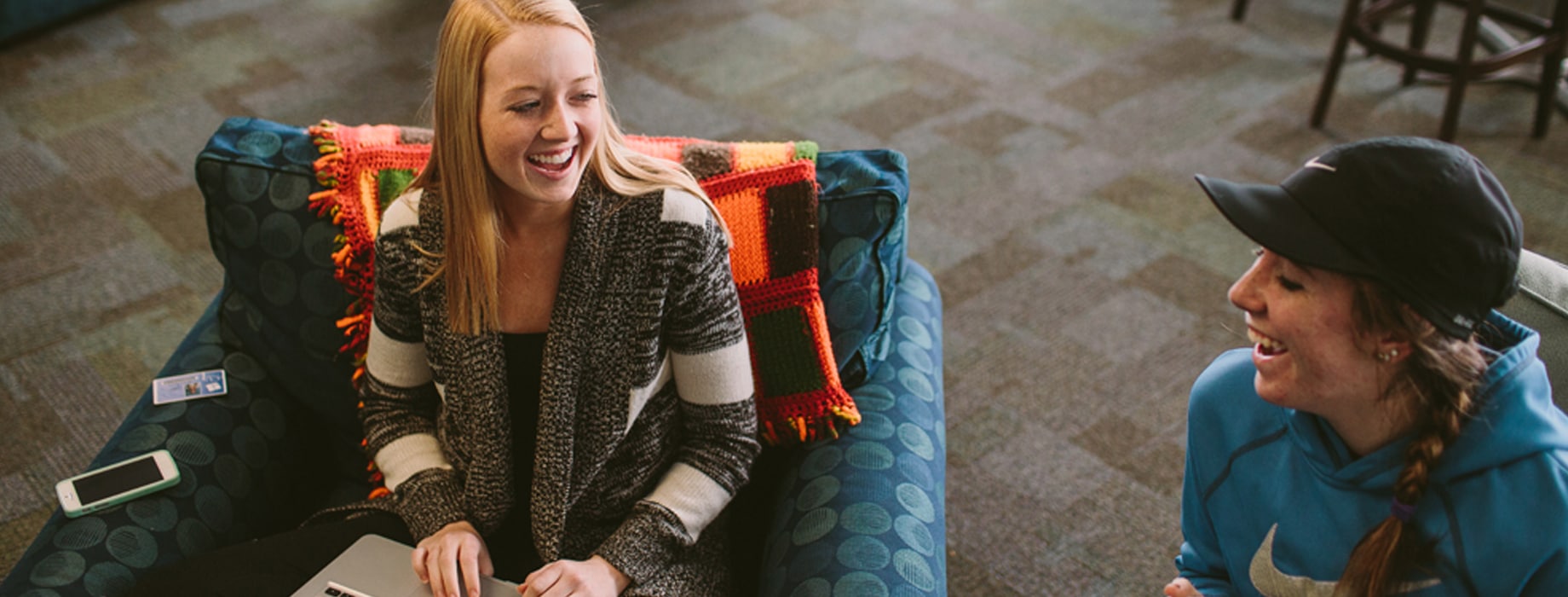 Two female college students sitting in chairs in dorm lobby while one holds a coffee mug and one types on a laptop computer