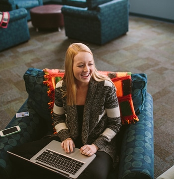 Two female college students sitting in chairs in dorm lobby while one holds a coffee mug and one types on a laptop computer