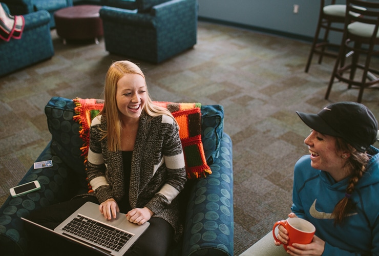 Two female college students sitting in chairs in dorm lobby while one holds a coffee mug and one types on a laptop computer