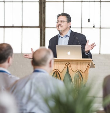 Adult male in a suit jacket standing at a wooden podium with a laptop while speaking to a room of guests