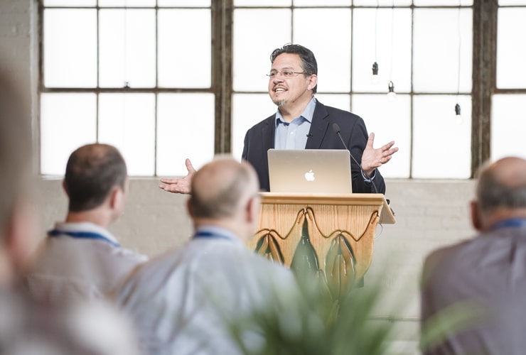 Adult male in a suit jacket standing at a wooden podium with a laptop while speaking to a room of guests