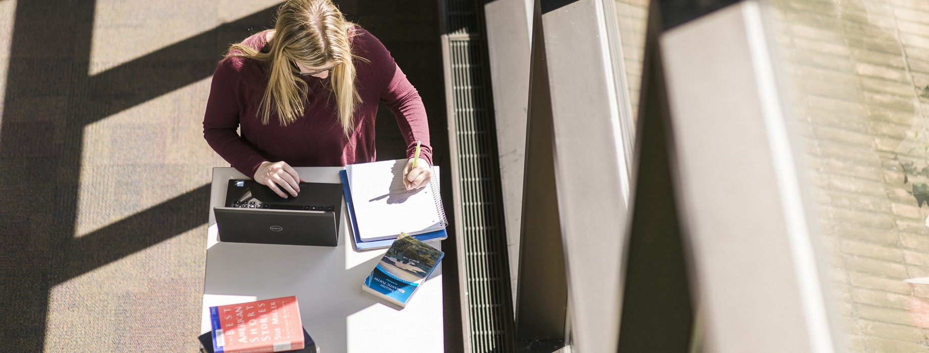 Female writing in notebook while standing at a desk with a laptop near a window in Miller Library at Cornerstone University