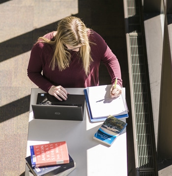 Female writing in notebook while standing at a desk with a laptop near a window in Miller Library at Cornerstone University