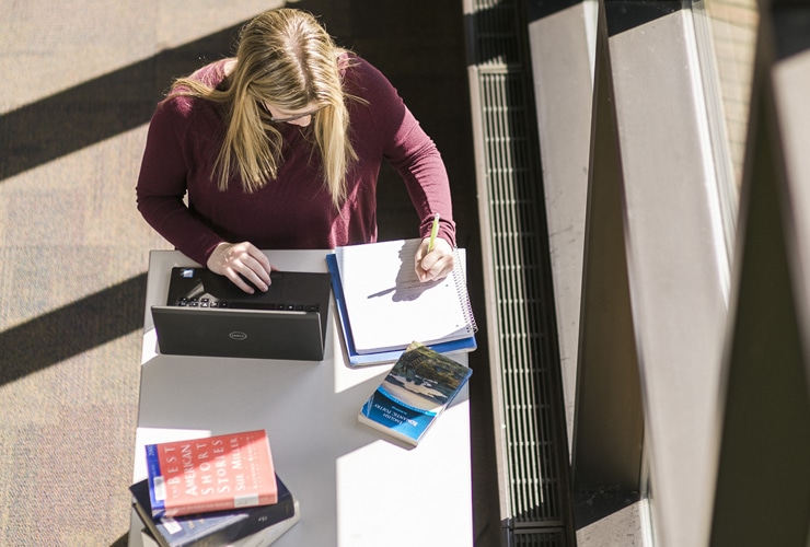 Female writing in notebook while standing at a desk with a laptop near a window in Miller Library at Cornerstone University
