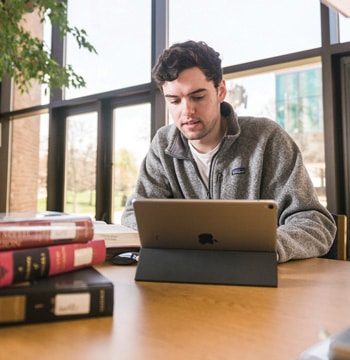 Male sitting at a wooden table in Miller Library at Cornerstone University with a tablet and stack of books