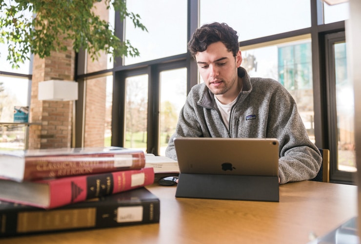 Male sitting at a wooden table in Miller Library at Cornerstone University with a tablet and stack of books