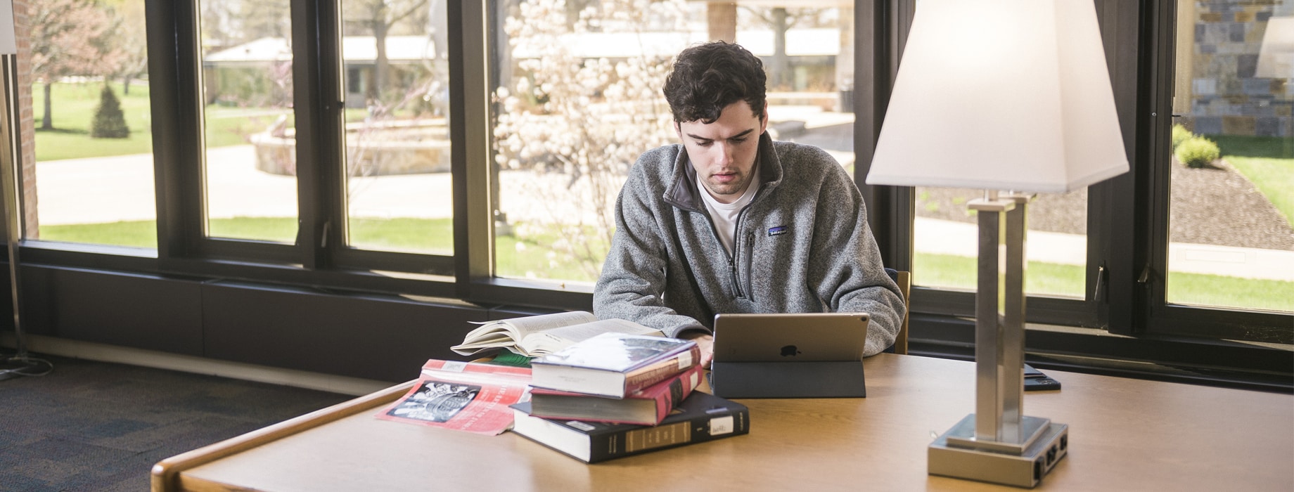Male typing on a tablet while sitting at a wooden table in Miller Library at Cornerstone University with a stack of books