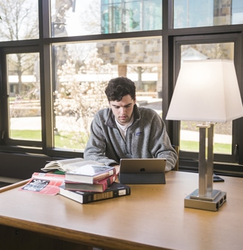 Male typing on a tablet while sitting at a wooden table in Miller Library at Cornerstone University with a stack of books