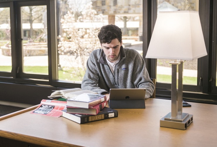 Male typing on a tablet while sitting at a wooden table in Miller Library at Cornerstone University with a stack of books