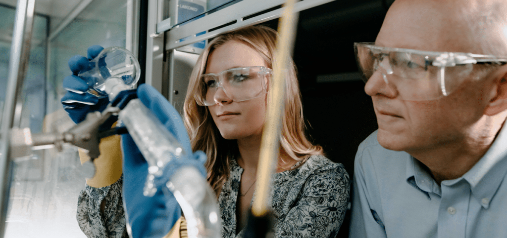 Female science student pouring a liquid from a beaker into a tube in a science lab at Cornerstone University under the guidance of a male professor